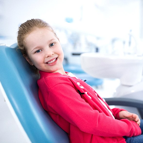 A little girl wearing a hot pink jacket sits in a dentist’s chair and smiles in preparation for her appointment