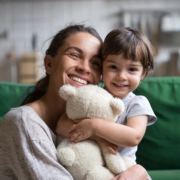 A mother hugs her daughter who holds onto a teddy bear and smiles