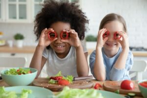Two girls holding hollowed out red peppers up as eyeglasses with salads nearby.